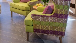 Un área de lounge moderna con sillones de brazos estampados en verde y morado, una pequeña mesa con un jarrón de flores y un aparador azul al fondo.