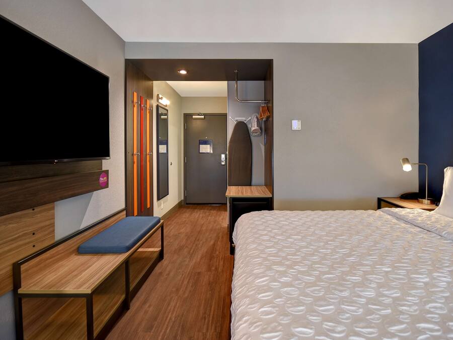 A hotel room with a bed, a bench, a TV, a desk, and a shower area.