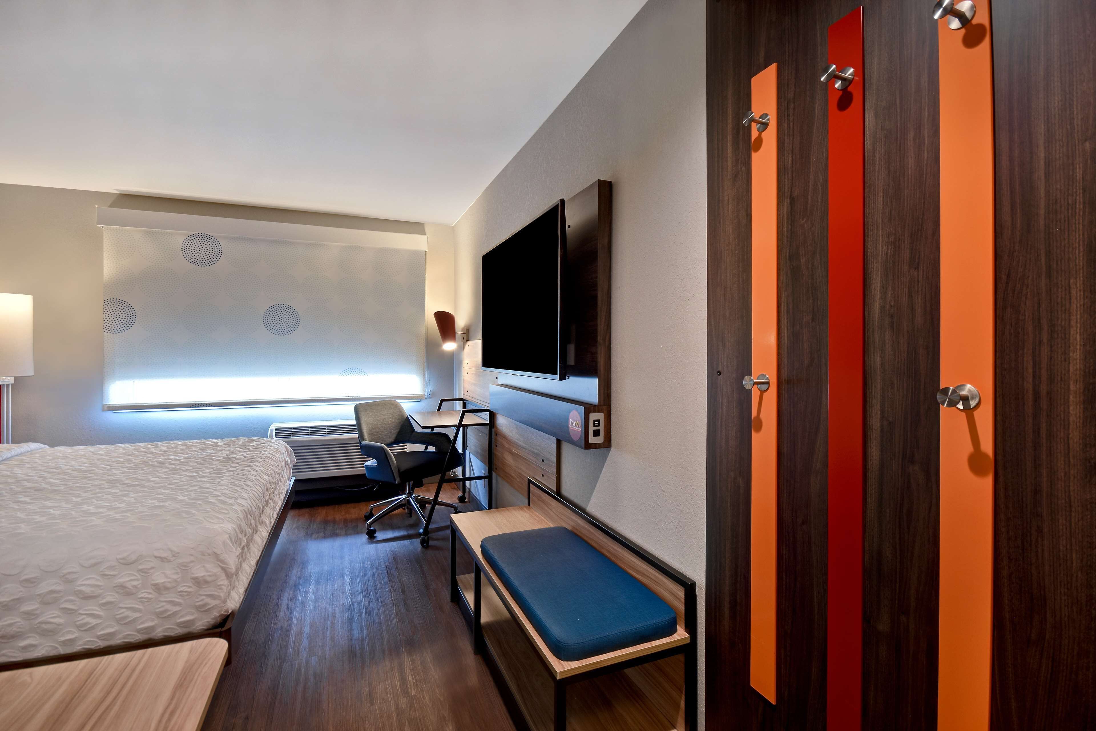 A hotel room with a bed, desk, chair, bench, TV, and a window with blinds.