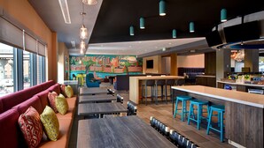 A modern restaurant with a long counter, blue bar stools, and a mural of Manchester. - Tru by Hilton Manchester Downtown (Manchester)