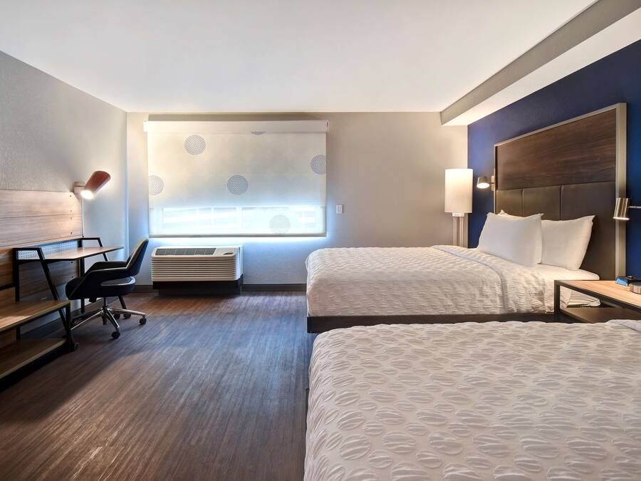 A hotel room with two beds, a desk, a chair, a TV, and a window with blinds.
