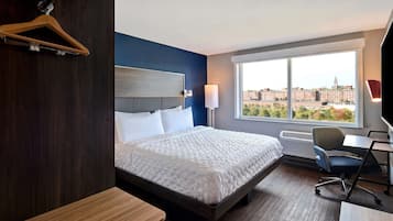 Ein Hotelzimmer mit einem großen Bett, einem Schreibtisch, einem Stuhl, einem Fenster mit Blick auf die Stadt und einem an der Wand befestigten Kleiderhaken.