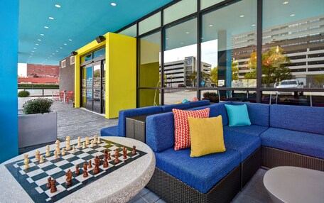 An outdoor seating area with a chess table, blue sofa with colorful cushions, and large windows overlooking a parking garage.
