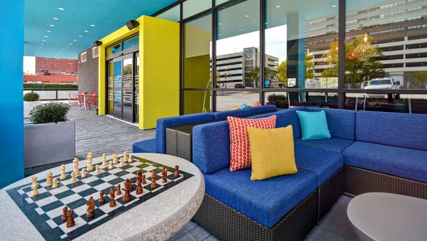 An outdoor seating area with a chess table, blue sofa with colorful cushions, and large windows overlooking a parking garage. - Tru by Hilton Manchester Downtown (Manchester)