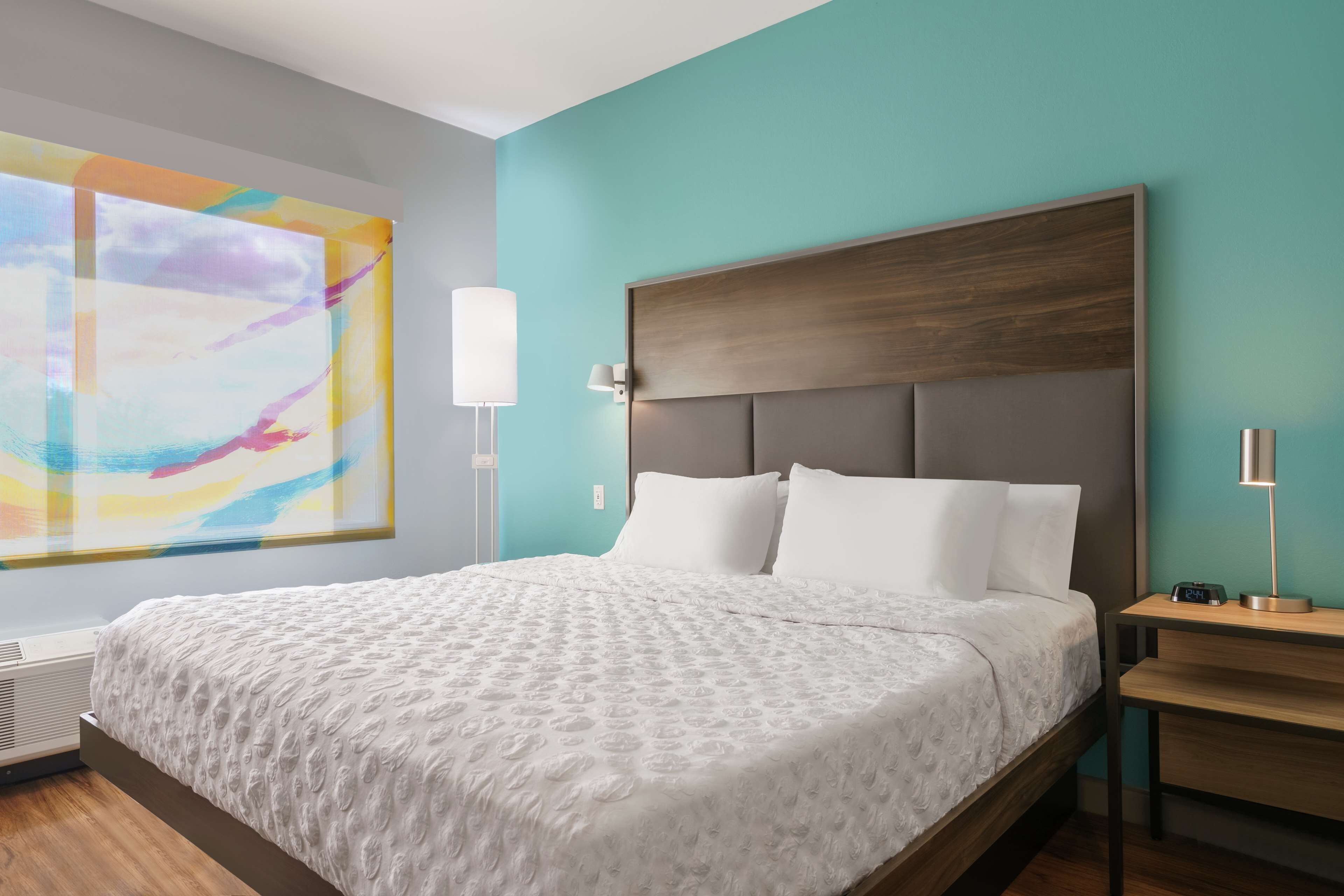 A modern hotel room with a large bed, a nightstand, a wall-mounted lamp, and a large abstract painting.