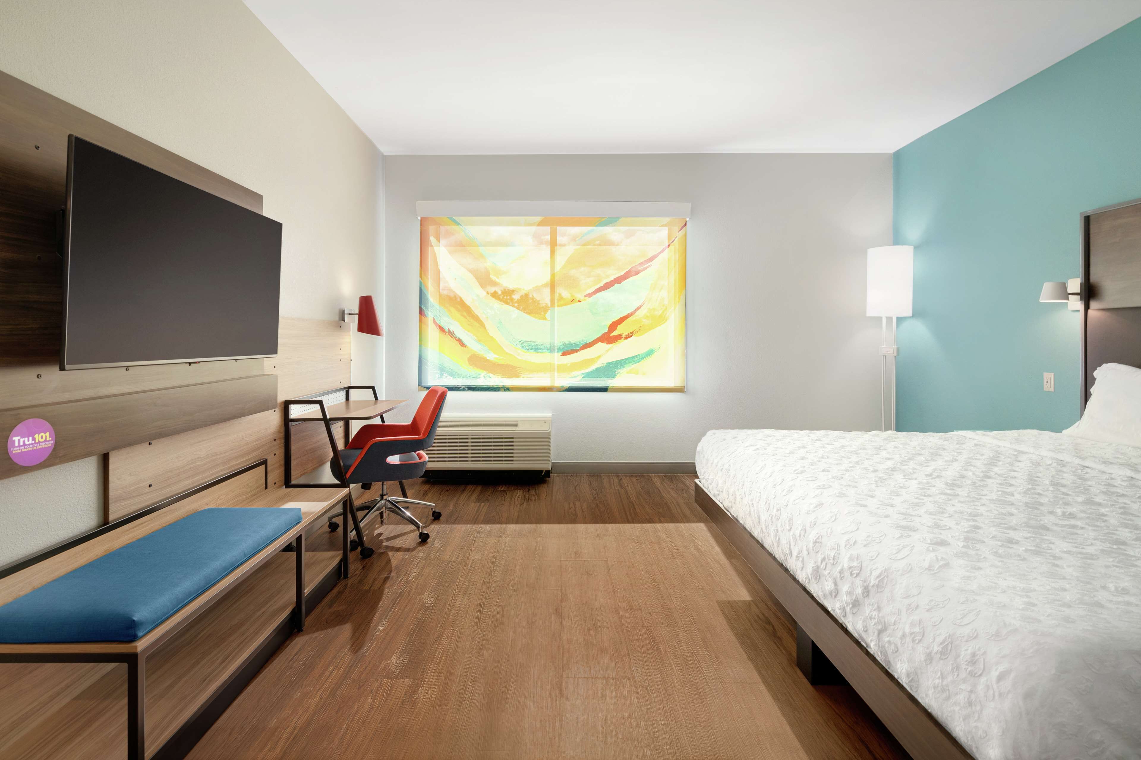 A modern hotel room with a bed, a desk, a chair, a flat-screen TV, a large window with a colorful abstract painting, and a wall-mounted lamp.