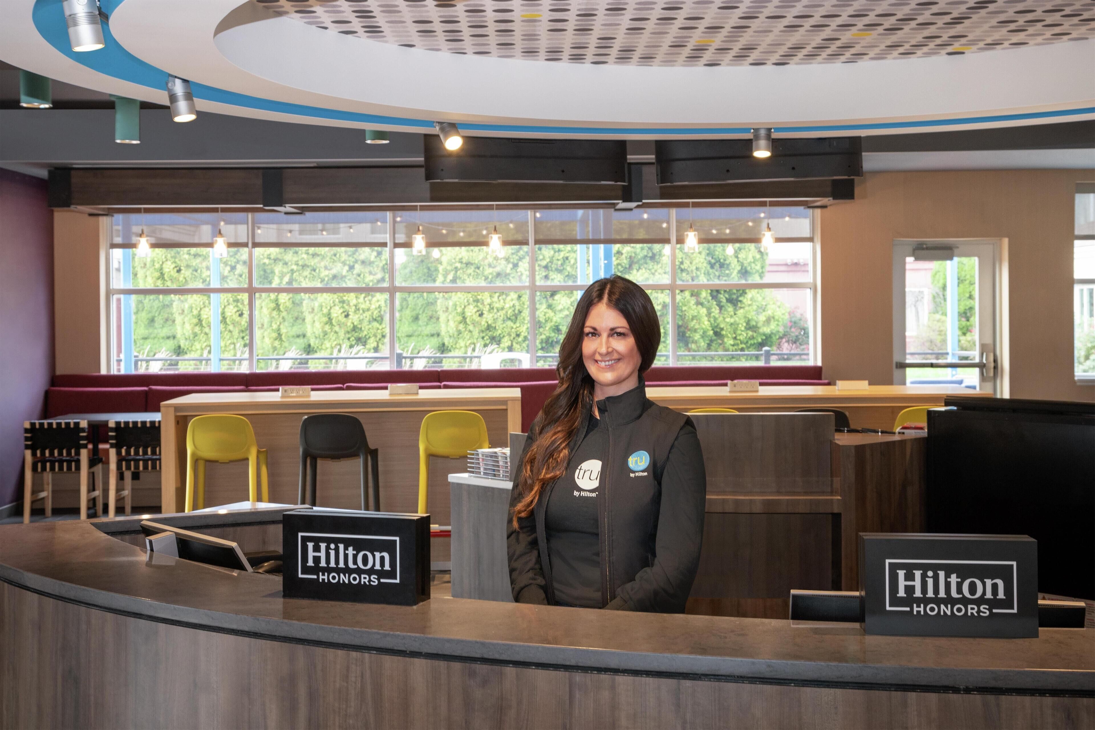 A reception desk with a "Hilton Honors" sign, a window with a view of greenery, and a seating area with yellow chairs.