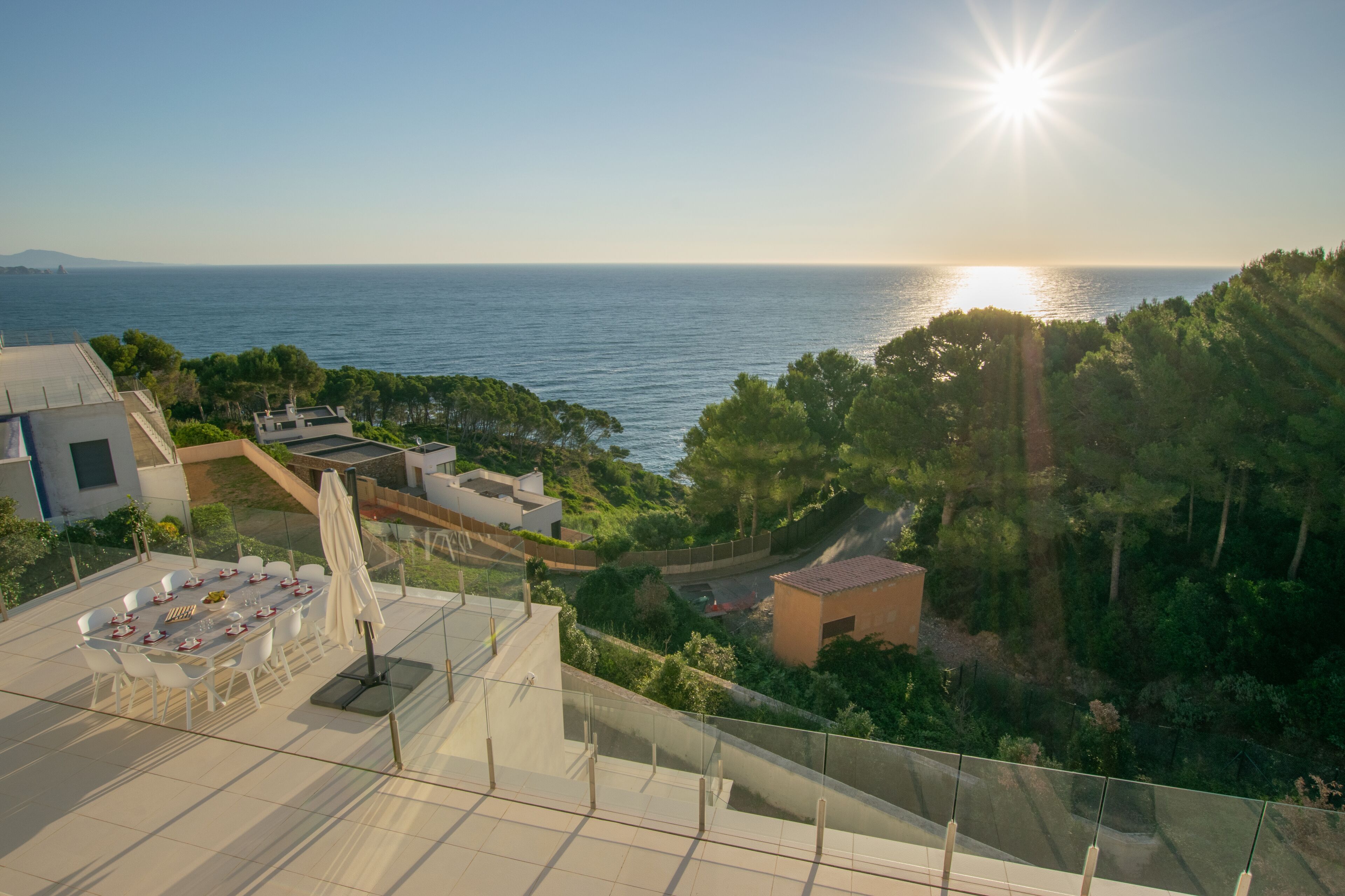 Eine Terrasse mit Esstisch und Stühlen, mit Blick auf eine Küstenlandschaft und freiem Blick auf das Meer und ferne Hügel.