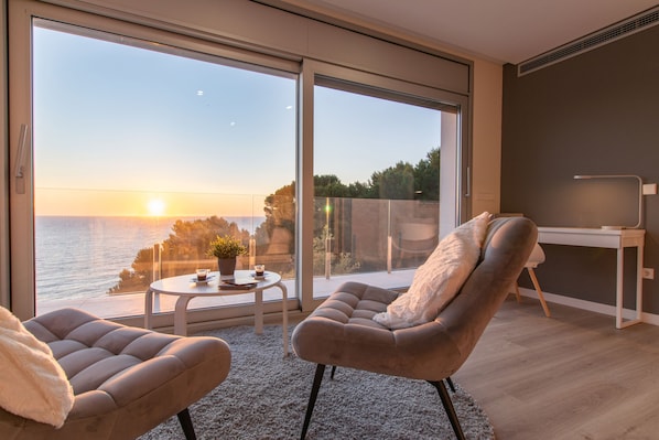 Een moderne kamer met een grote schuifglazen deur die leidt naar een balkon met uitzicht op een ondergaande zon boven het water. Er staat een ronde tafel met een plant, een bank en een bureau met een stoel.