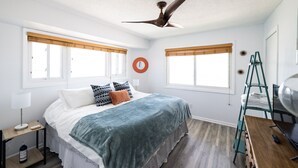 Quarto com cama, duas janelas com persianas, ventilador de teto, mesa de cabeceira com luminária e escada.