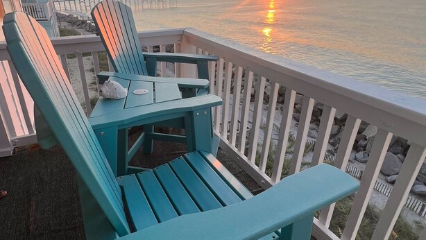Uma cadeira Adirondack azul-esverdeada numa varanda com vista para o pôr do sol sobre a água.