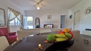 A dining area with a table set for four, a television in the background, and a view of the outdoors through a window. - Herdade Palma T1  (country house) (Moita)