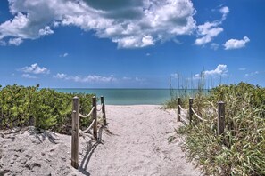 Een zandpad langs het strand dat naar de oceaan leidt, omringd door groen en houten palen.