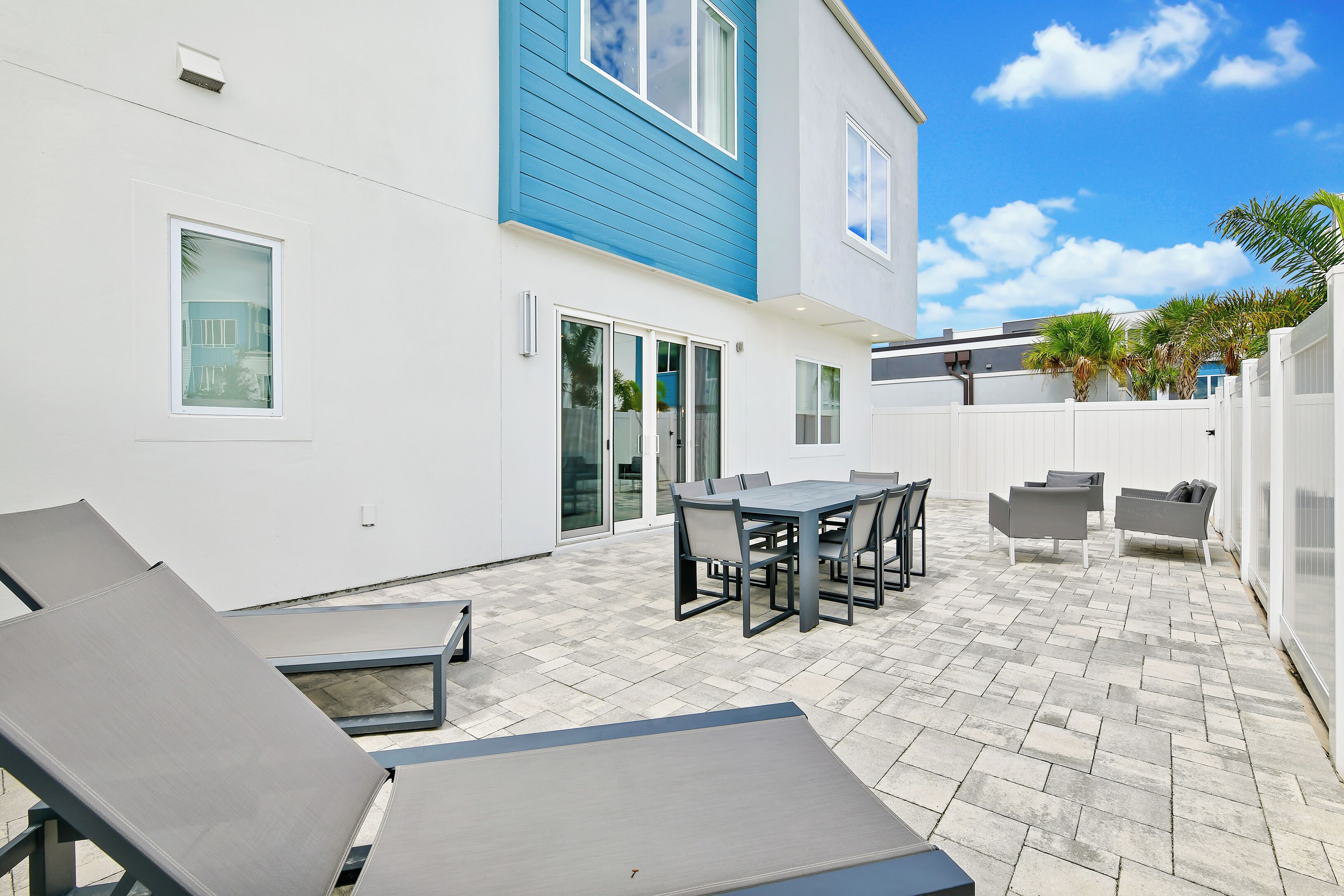 A modern outdoor patio with a dining table and chairs, a lounge area with sun loungers, and a white building with blue trim.