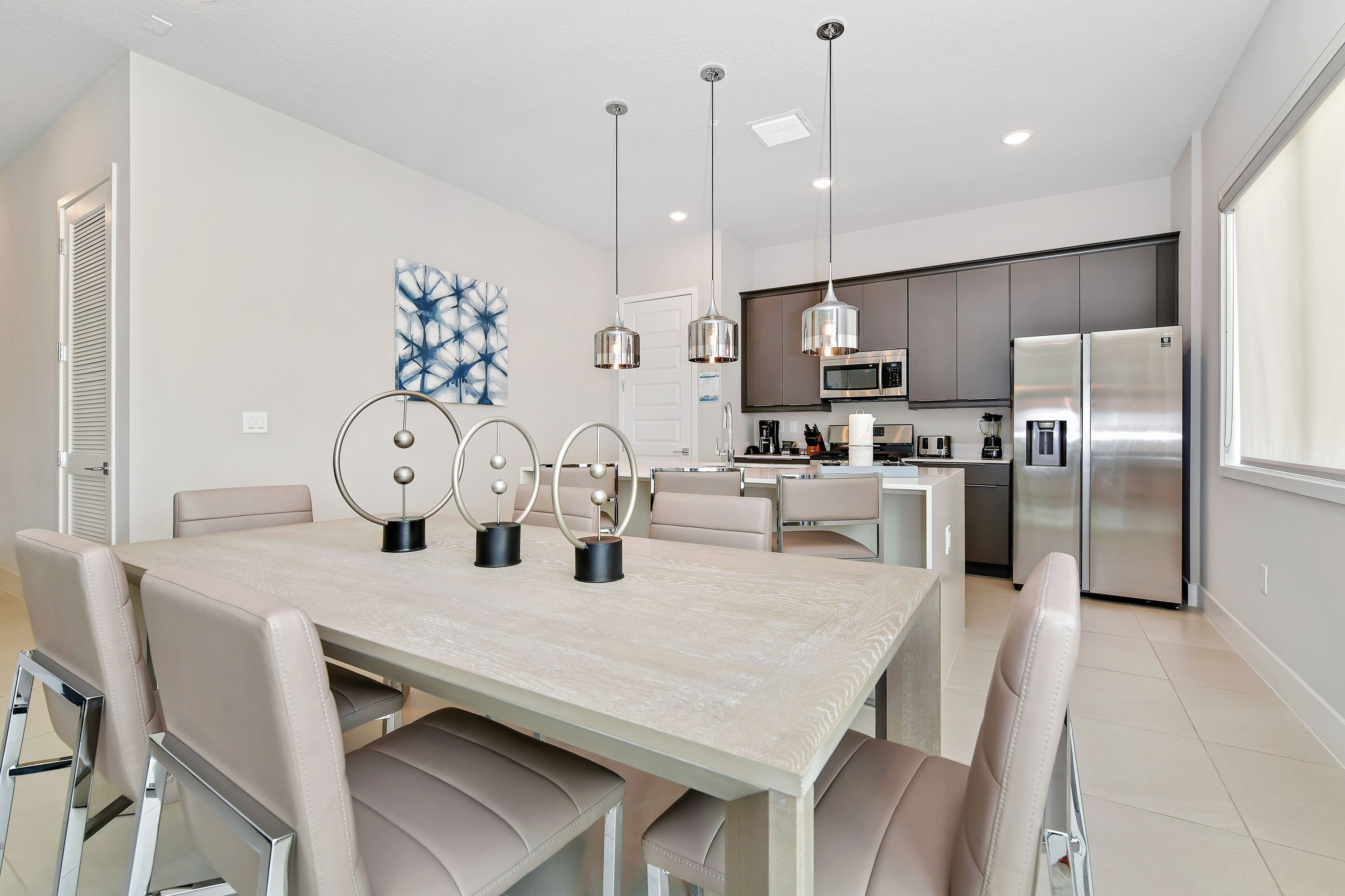 Een moderne keuken met roestvrijstalen apparatuur, een eethoek met een licht houten tafel en beige stoelen, en hanglampen.