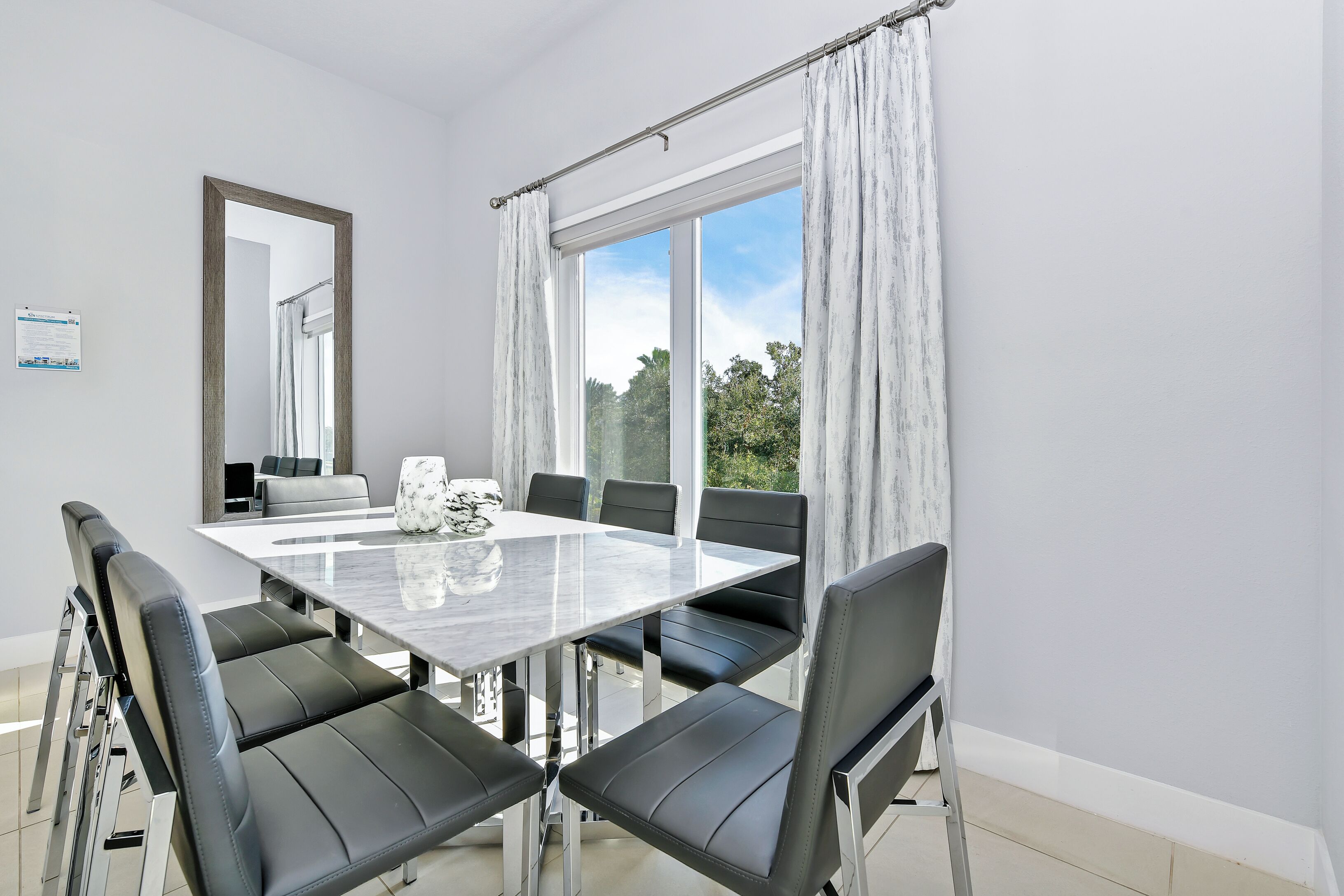 Un comedor moderno con una mesa de vidrio y sillas de cuero negro, un espejo grande y vistas a la vegetación exterior.