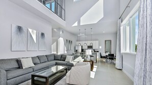 A modern living room with a sectional sofa, a glass coffee table, and a white armchair. There is a kitchen area in the background with pendant lighting. - Spectrum Resort Villas Orlando and Waterparks (Kissimmee)