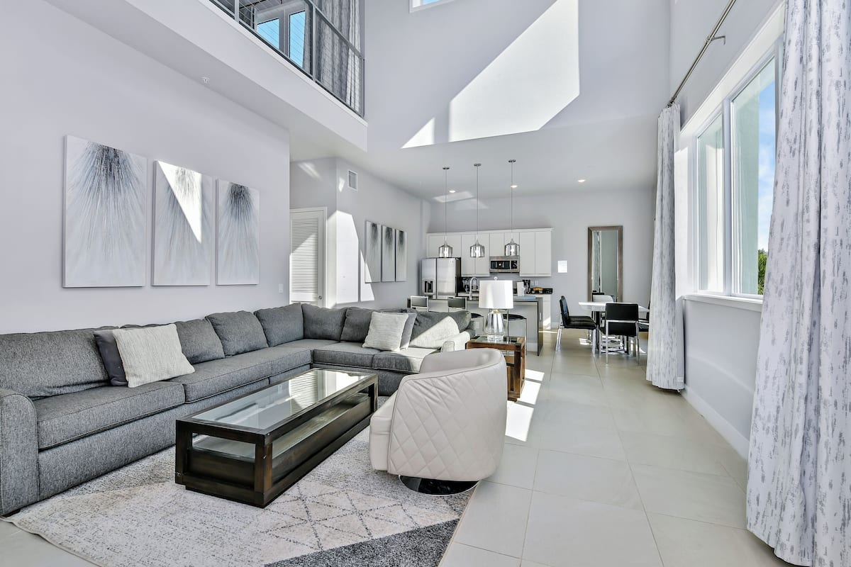 A modern living room with a sectional sofa, a glass coffee table, and a white armchair. There is a kitchen area in the background with pendant lighting.