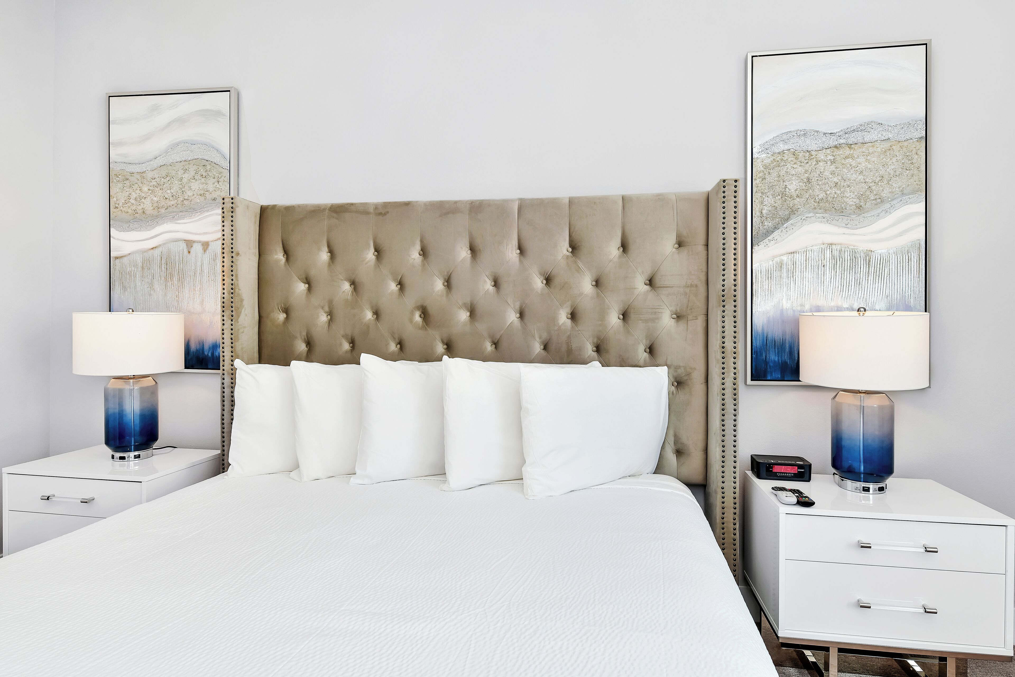 A hotel room with a tufted headboard, white bedding, two bedside tables with lamps, and artwork on the walls.