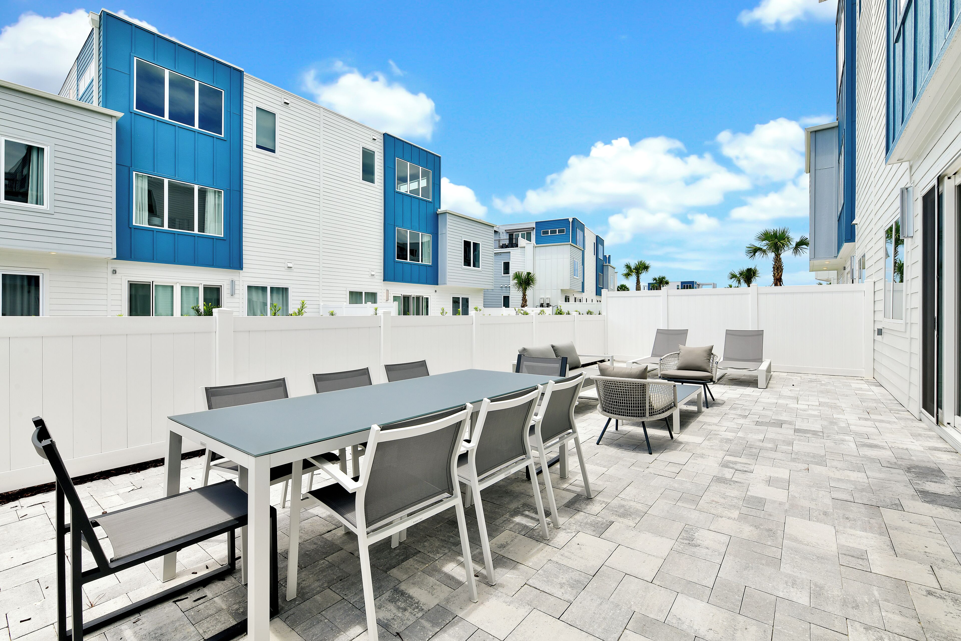 A patio with a table and chairs, a white fence, and a blue building in the background.