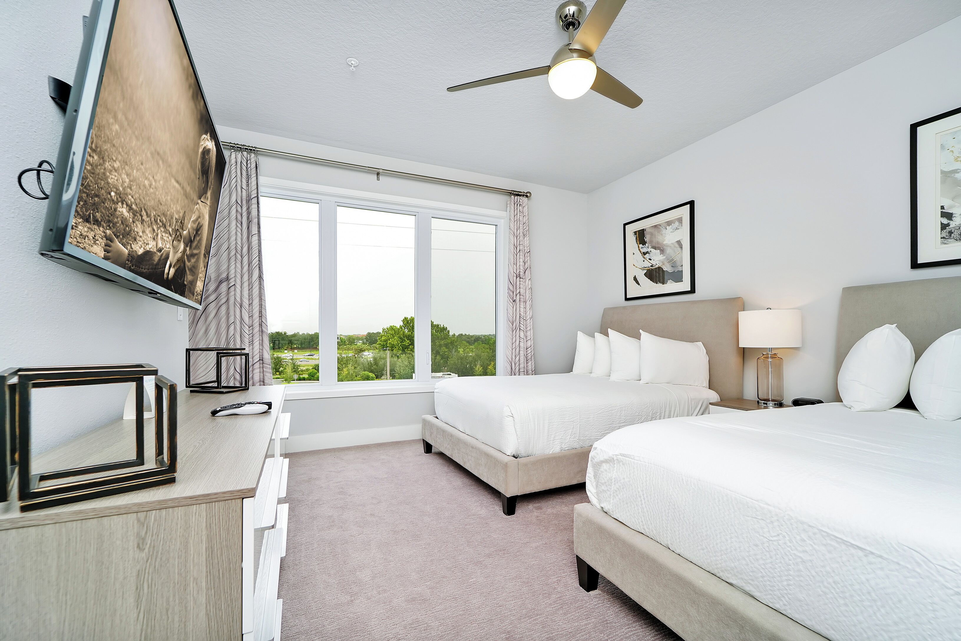 A hotel room with two beds, a ceiling fan, a large window, and a TV mounted on the wall.