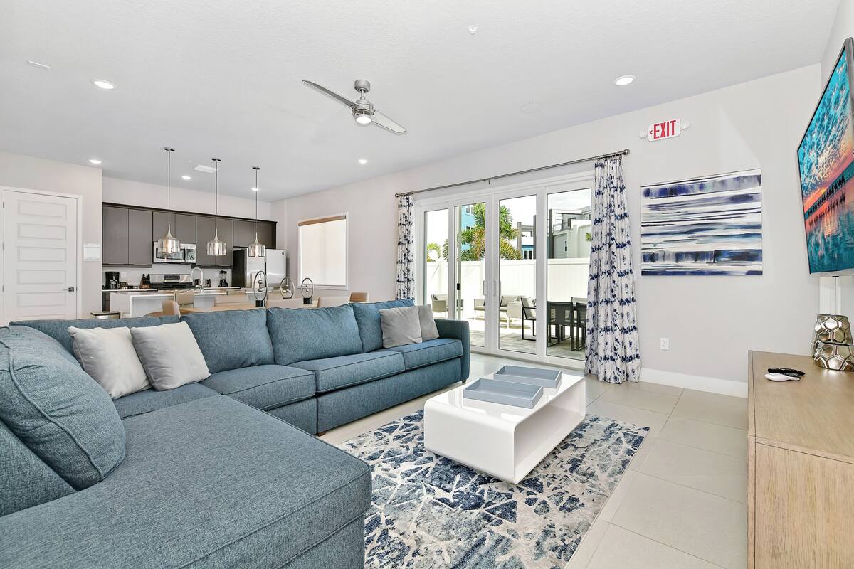 A modern living room with a sectional sofa, a coffee table, and a view of the kitchen and dining area through large sliding glass doors.