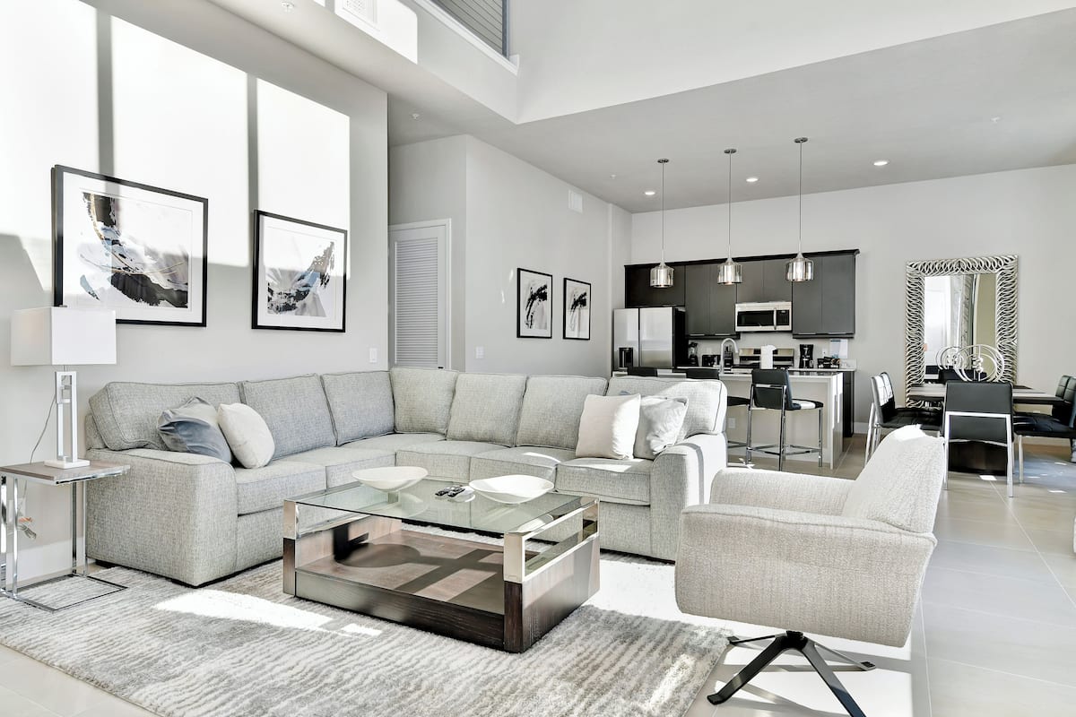 A modern living room with a grey sofa, a glass coffee table, and a dining area with a kitchen in the background.