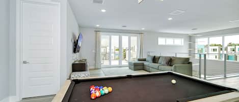 A pool table with balls, a sofa, and a TV in a modern room with large windows.