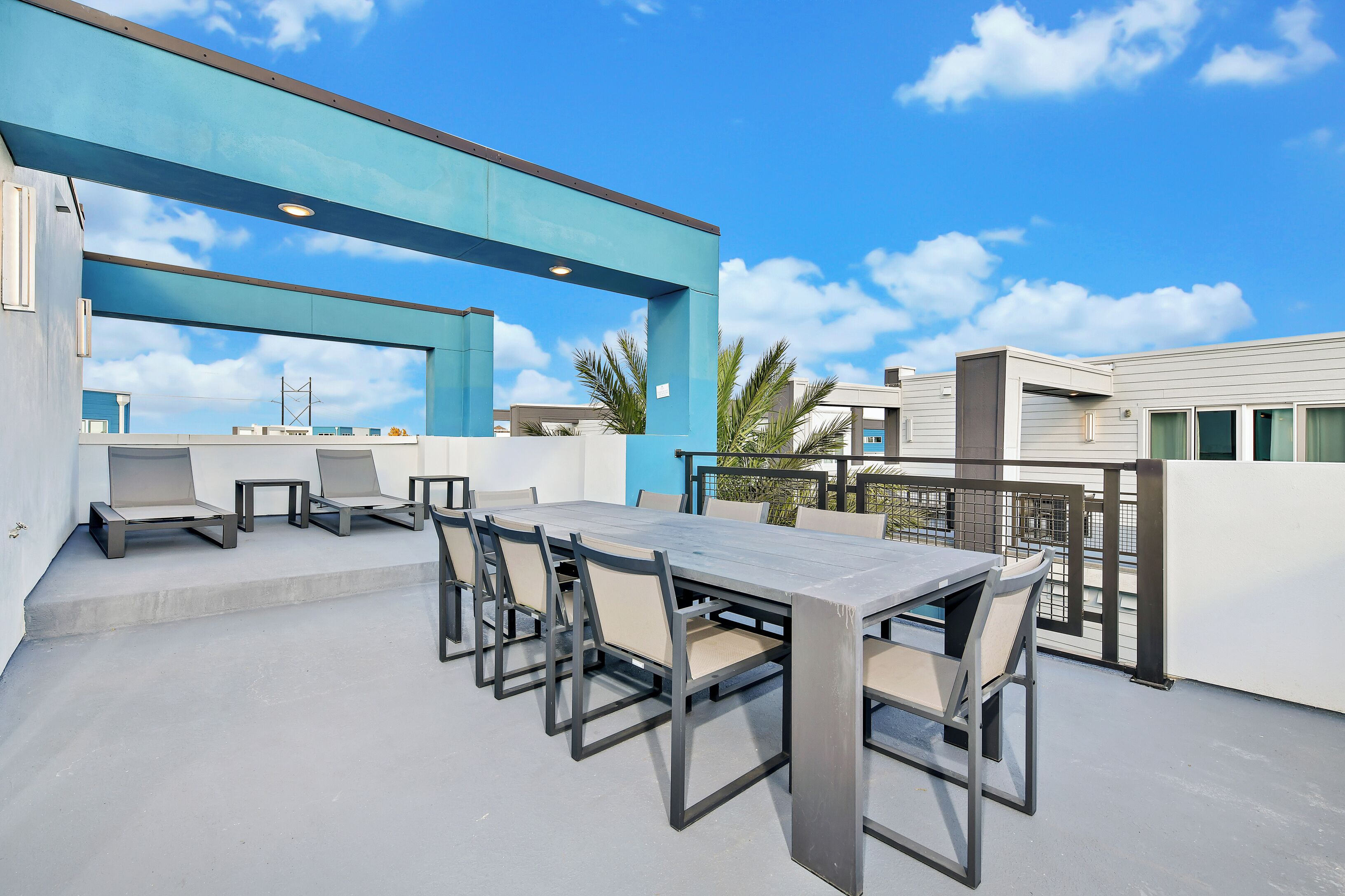 An outdoor patio with a long table and chairs, a blue overhang, and a view of palm trees and buildings.