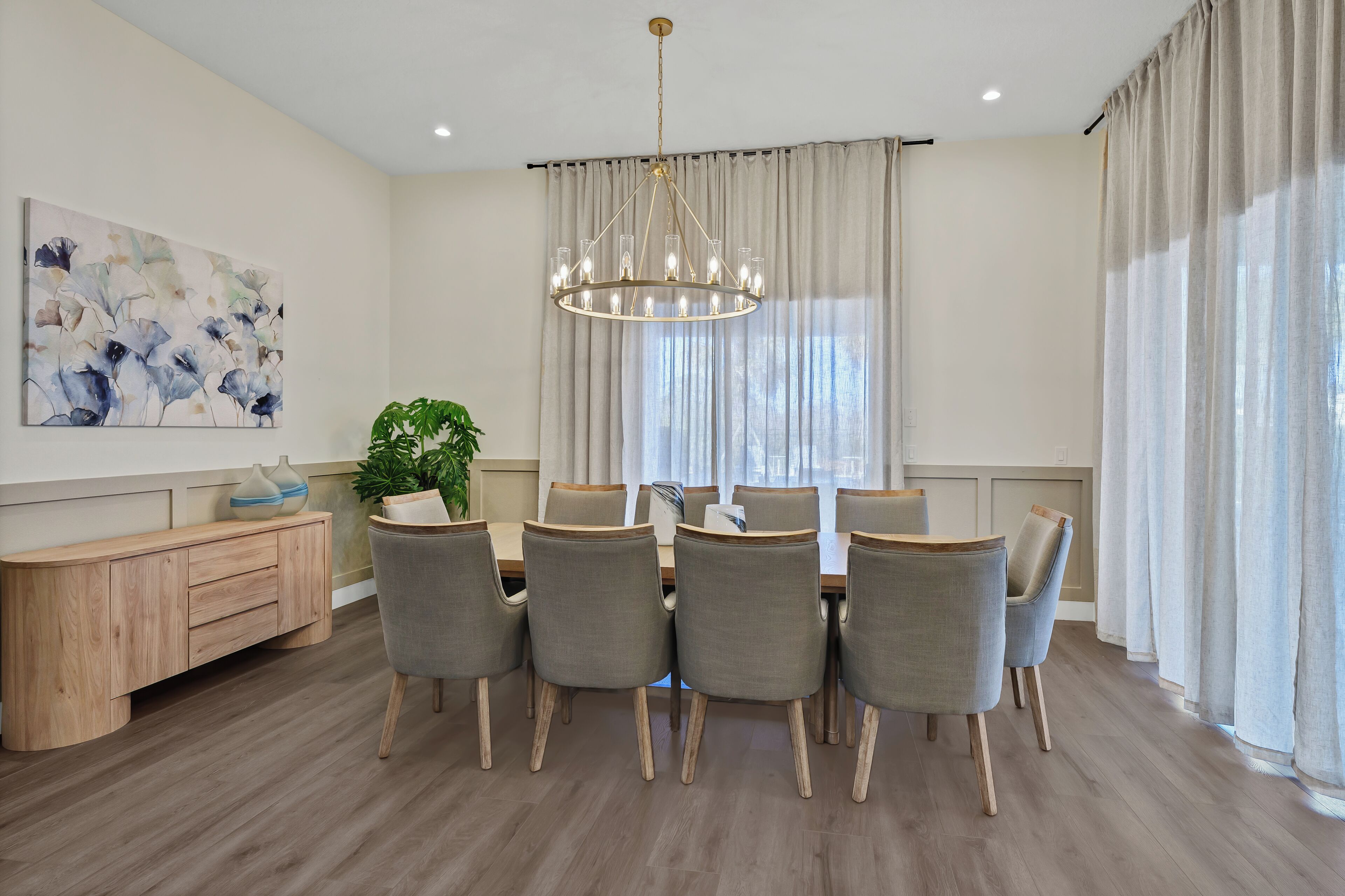 A dining area with a wooden table, six chairs, a chandelier, a plant, and a painting on the wall.