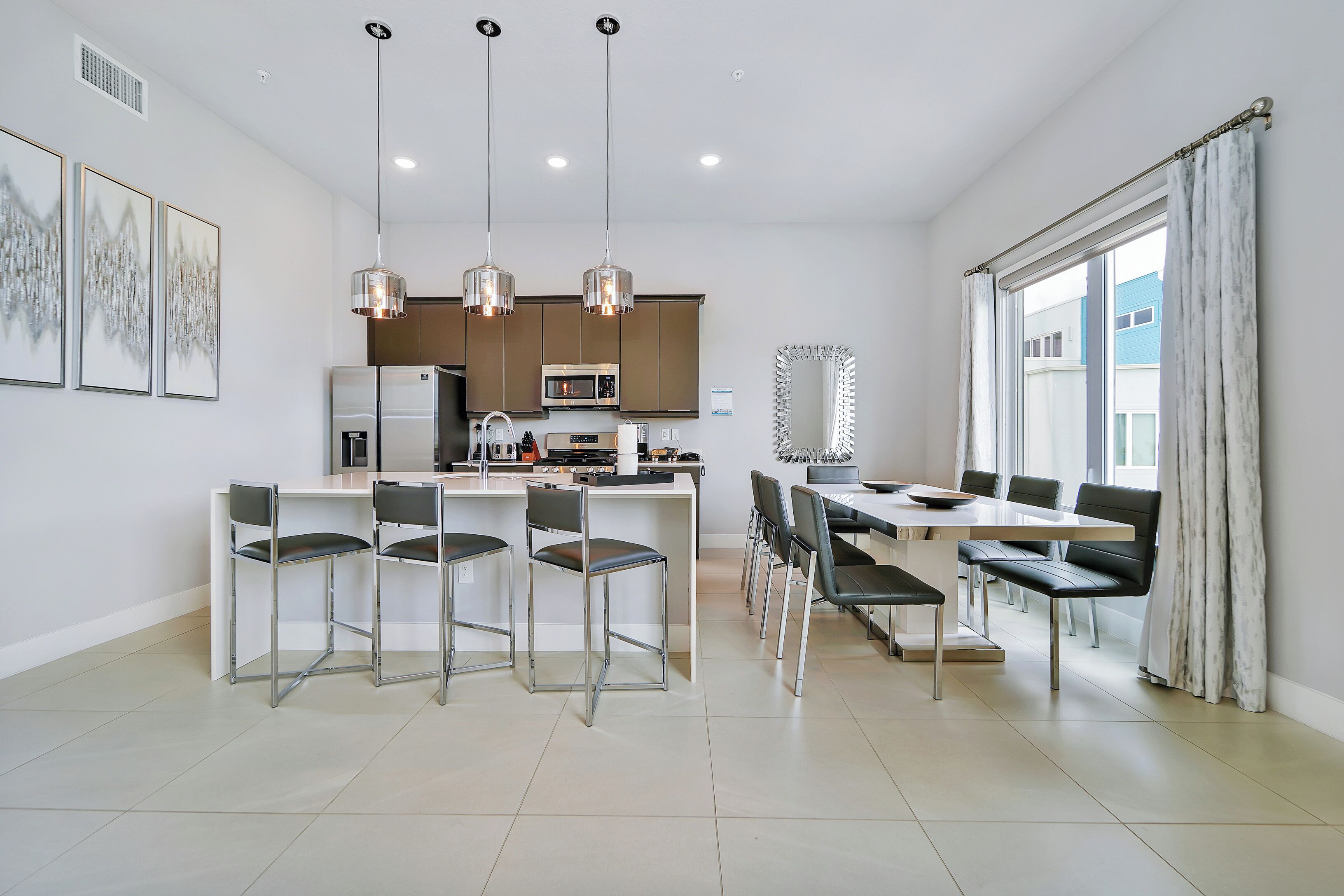 Una cocina moderna con electrodomésticos de acero inoxidable, un comedor con mesa y sillas, y ventanas amplias con cortinas.