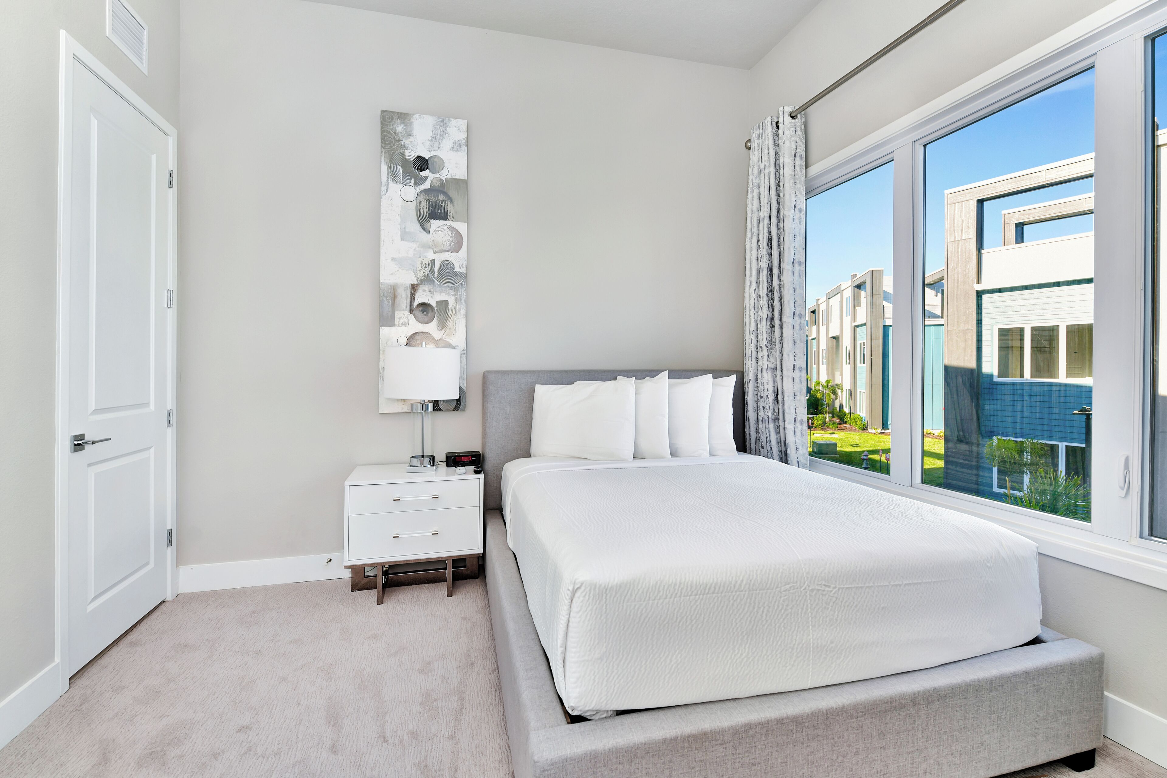 A bedroom with a large bed, a nightstand, a wall-mounted lamp, and a window with a view of buildings.