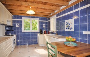 Une cuisine avec des carreaux bleus, des poutres en bois au plafond, une fenêtre et une table à manger sur laquelle se trouve un panier de fruits.