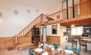 Un espace repas avec un escalier en bois, une bibliothèque et, au fond, une cuisine.