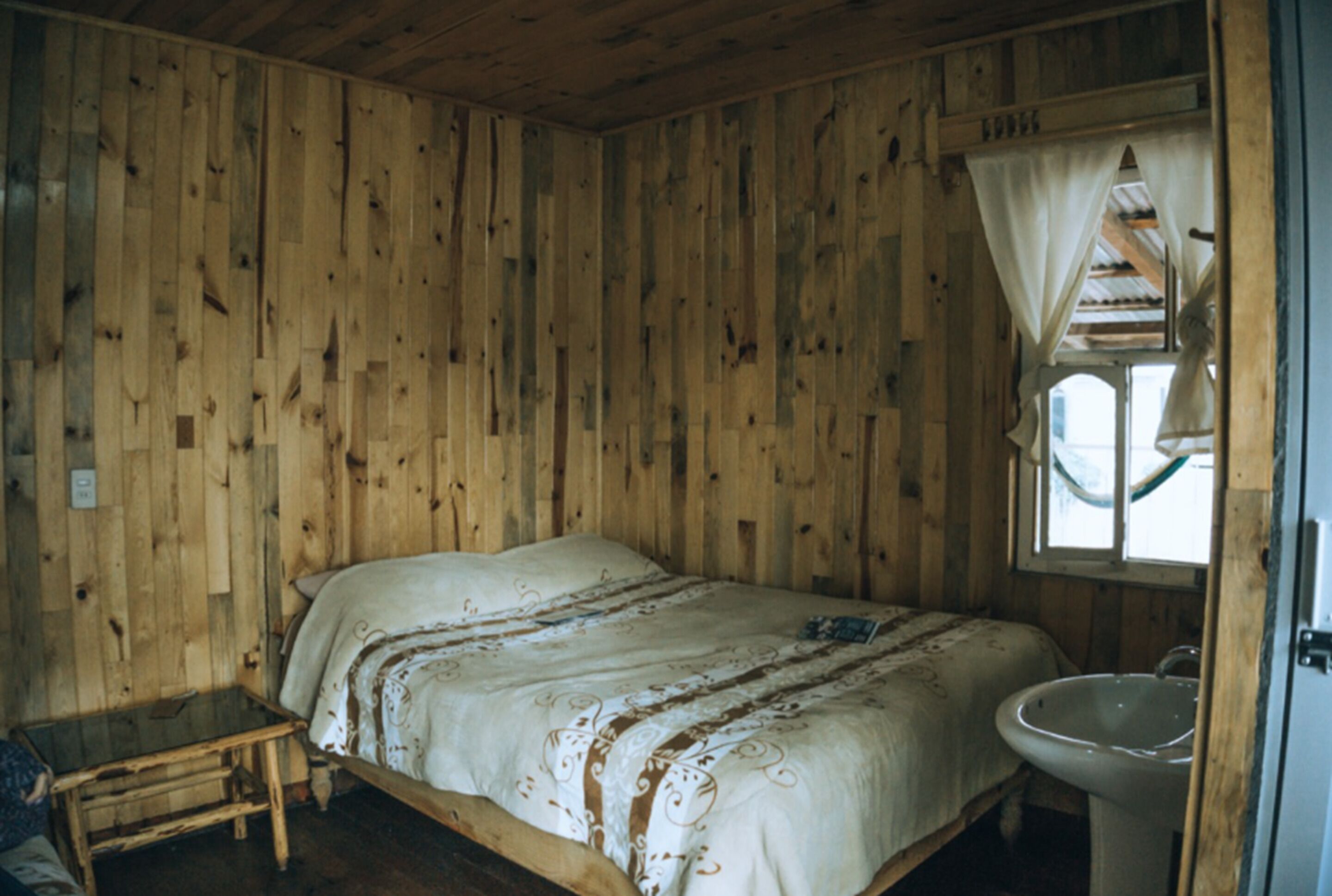 Una habitación de madera con una cama, una mesita y una ventana con cortinas.