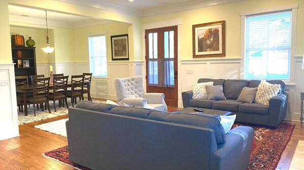 A living room with a grey sofa, a dark grey couch, and a wooden dining table with chairs. There is a chandelier hanging from the ceiling, a framed picture on the wall, and a window with blinds. - Beautiful Cottage on Hilton Head Island, Blue (Hilton Head Island)