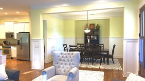 A modern kitchen with built-in appliances, a dining area with a wooden table and chairs, and a seating area with a tufted armchair. - Beautiful Cottage on Hilton Head Island, Blue (Hilton Head Island)