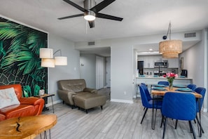 Ein modernes Wohnzimmer mit Deckenventilator, ein Essbereich mit blauen Stühlen und eine Küche im Hintergrund.