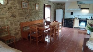 A cozy living room with a stone wall, a wooden dining table with chairs, a sofa, and a kitchen area with cabinets and appliances. - Rural house (pet-friendly) where you will only listen to nature. (Rianxo, A Coruña)