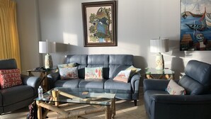 Een woonkamer met een blauwe bank, een glazen salontafel en twee bijzettafels met lampen. Er hangen twee kunstwerken in lijsten aan de muur.
