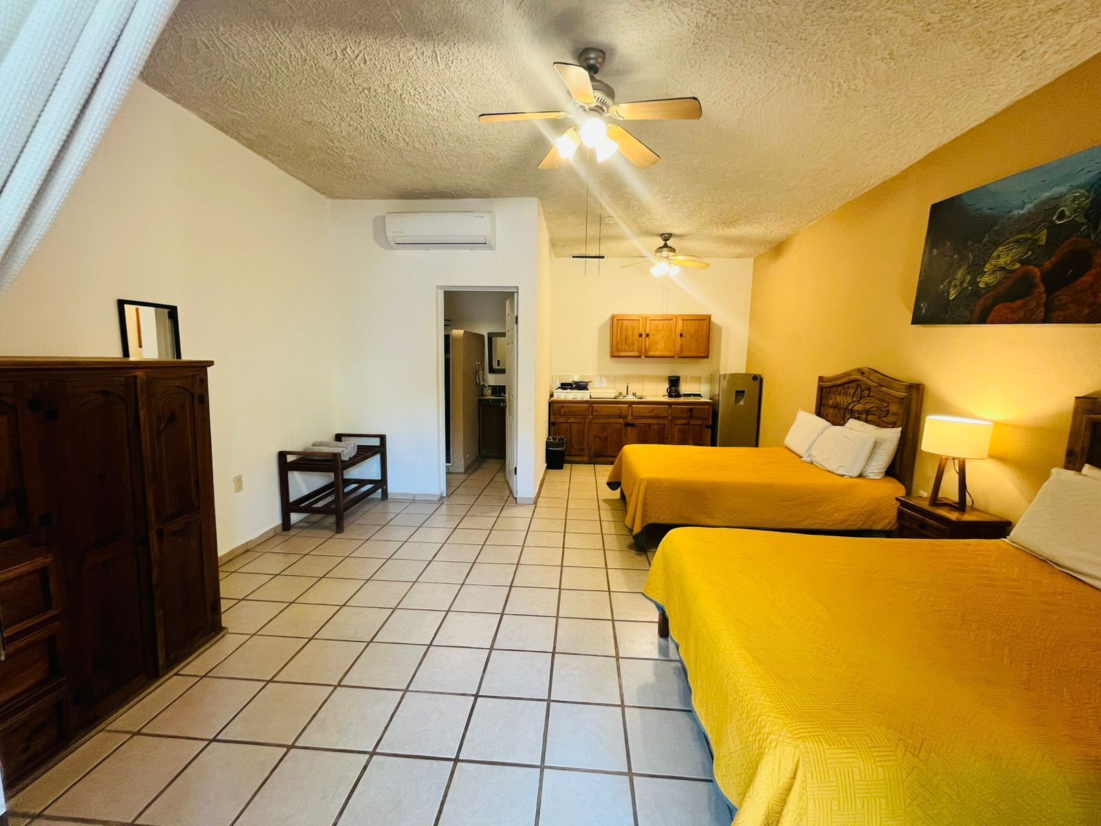 A hotel room with two beds, a kitchenette, a ceiling fan, and a painting on the wall.