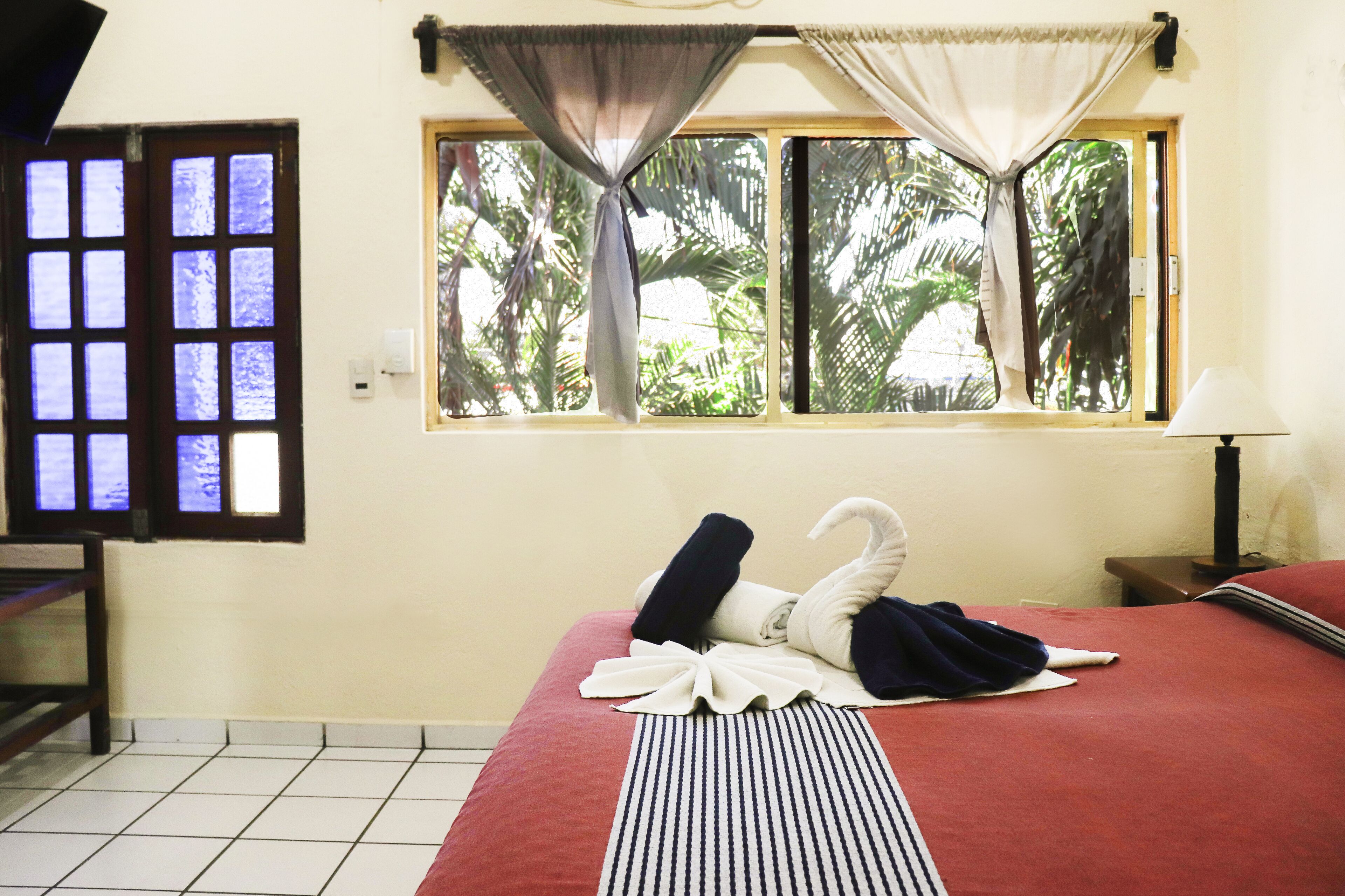 A hotel room with a bed, a window with curtains, a bedside table with a lamp, and a view of palm trees outside.