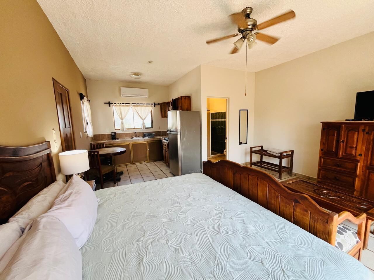 A bedroom with a bed, a ceiling fan, a wooden dresser, a desk, and a kitchenette in the background.