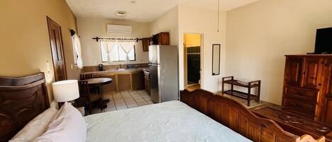 Quarto com cama, ventoinha no teto, cómoda de madeira, escrivaninha e uma kitchenette ao fundo.
