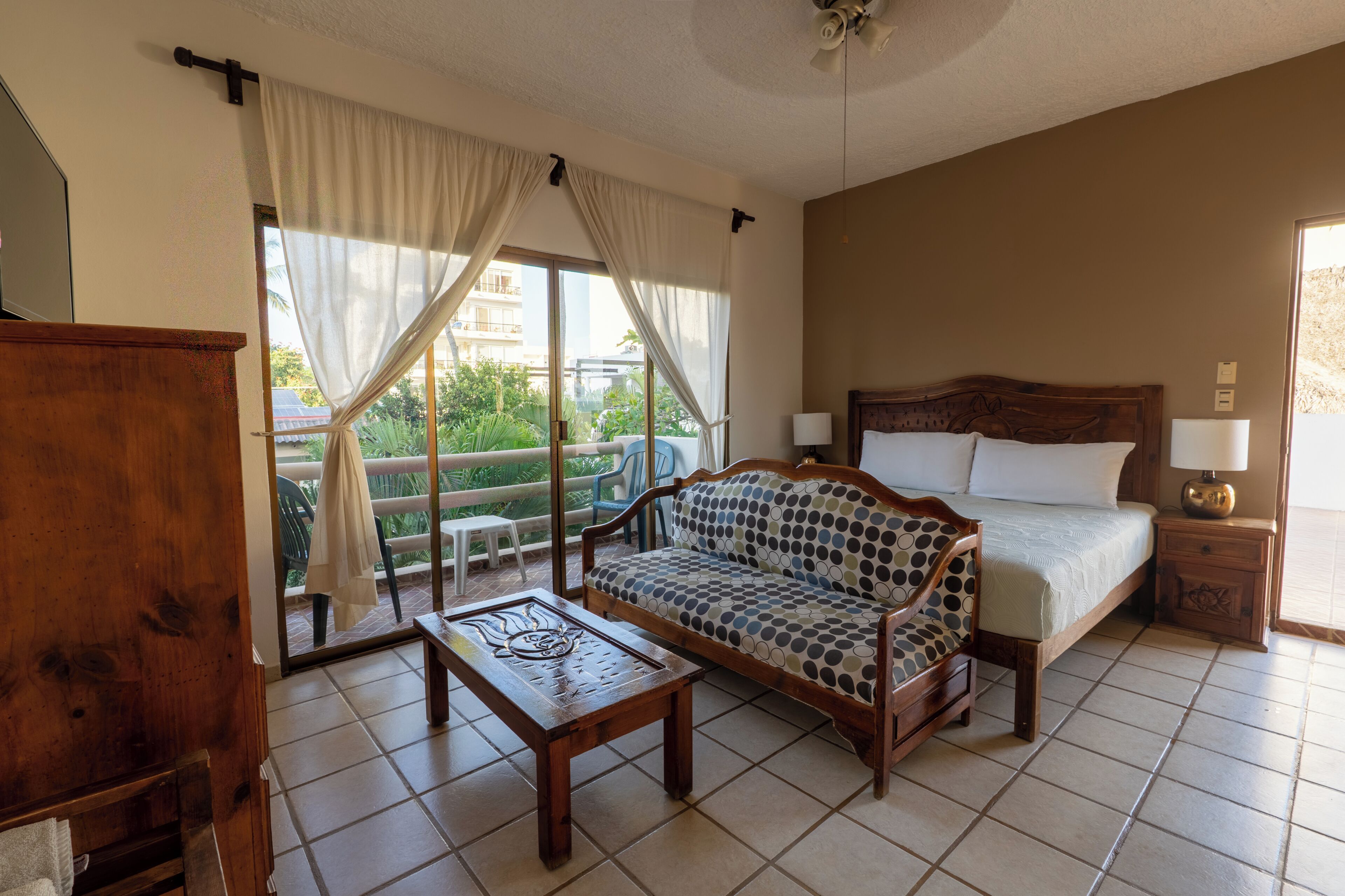 A hotel room with a bed, a sofa, a wooden table, and a balcony with a view of greenery.