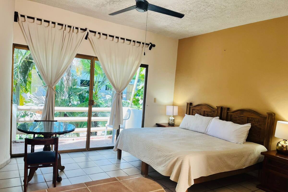 Habitación de hotel con cama, mesita de noche, mesa con silla y vistas a un balcón con cortinas.