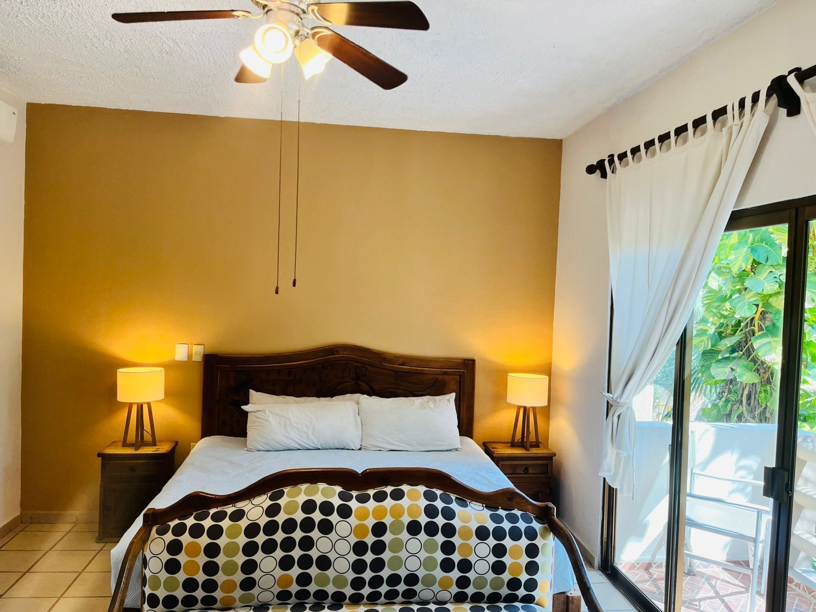 A bedroom with a bed, two bedside lamps, a ceiling fan, and a view of greenery through a window.