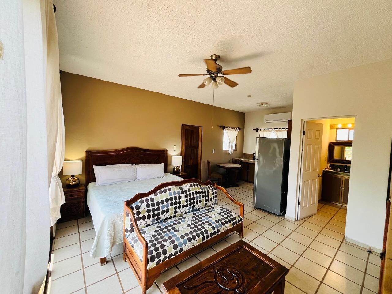 A hotel room with a bed, a sofa, a coffee table, a ceiling fan, a kitchenette, and a bathroom.