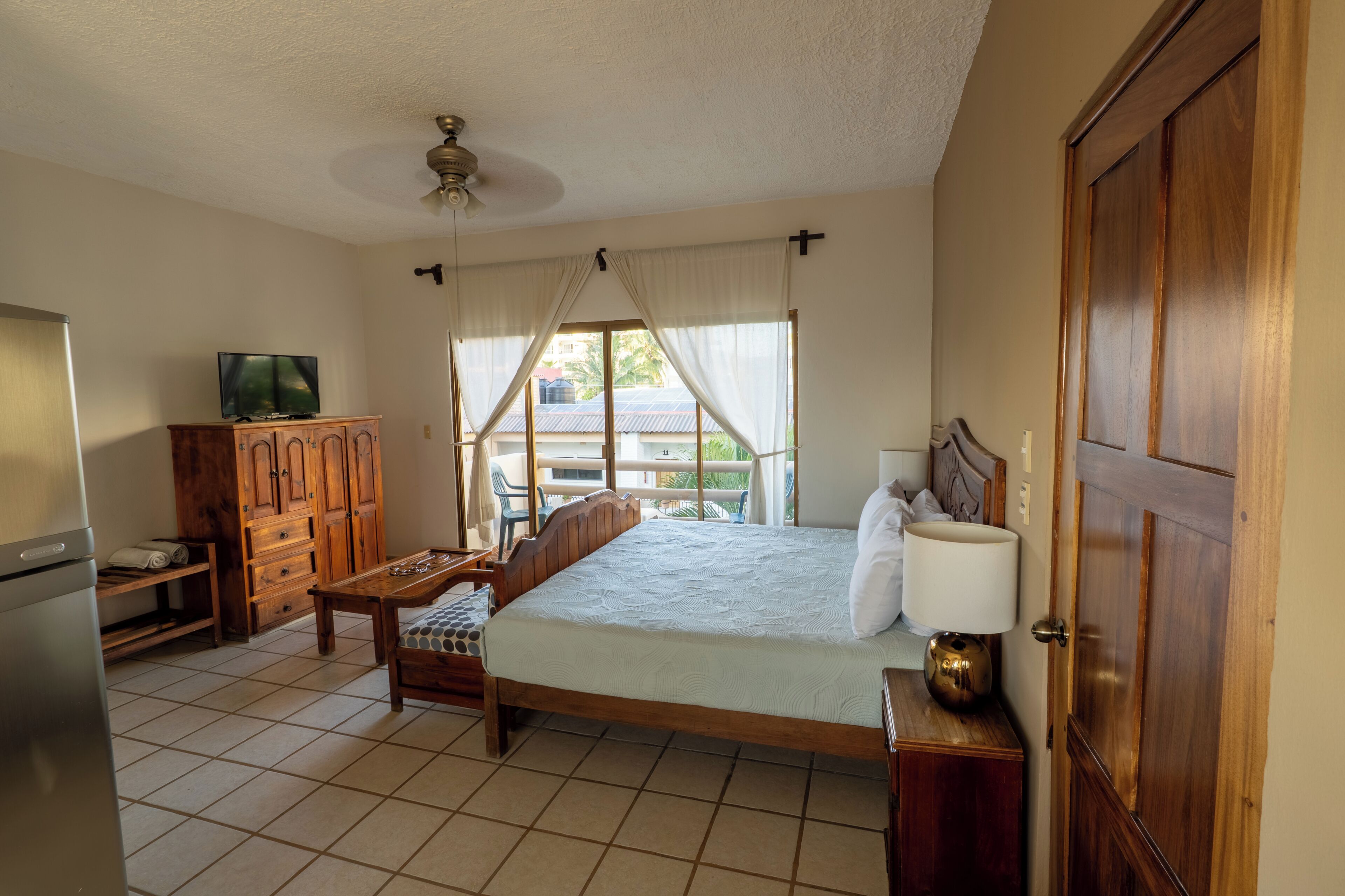 Un dormitorio con cama, televisor, armario de madera y ventana con cortinas.