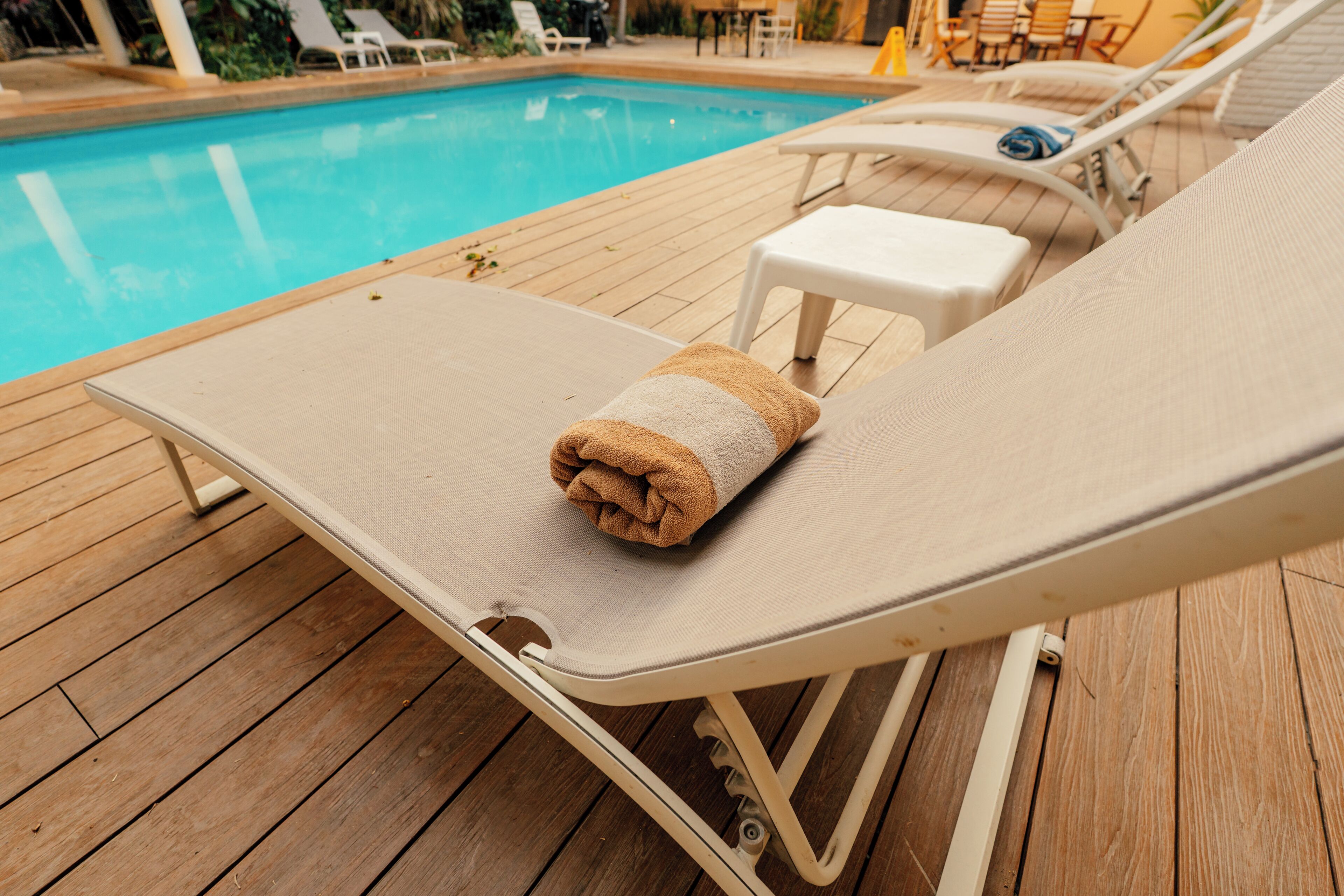 Una silla de descanso junto a la piscina, con una toalla, terraza de madera y piscina al fondo.
